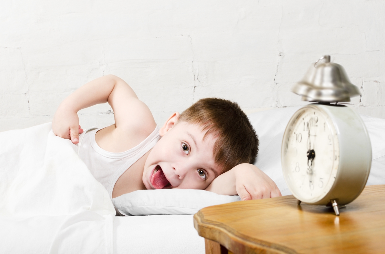 Small toddler boy (4 years old) is lying in bed and showing tongue. He is looking at the camera. Old clock show 6 o'clock. Brick wall